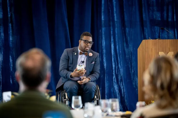 Dr. Feranmi Okanlami, Director of Student Accessibility and Accommodation Services (SAAS), on a stage speaking to a group. He has brown skin, black hair, black glasses, a blue suit with a maroon bow tie, and is in a wheelchair.