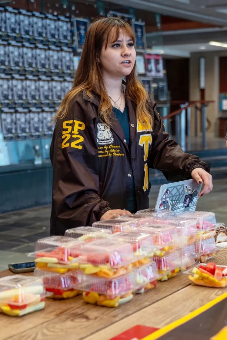 Close-up of a student in a lettered jacket smiling behind a table filled with to-go packages of fruit.