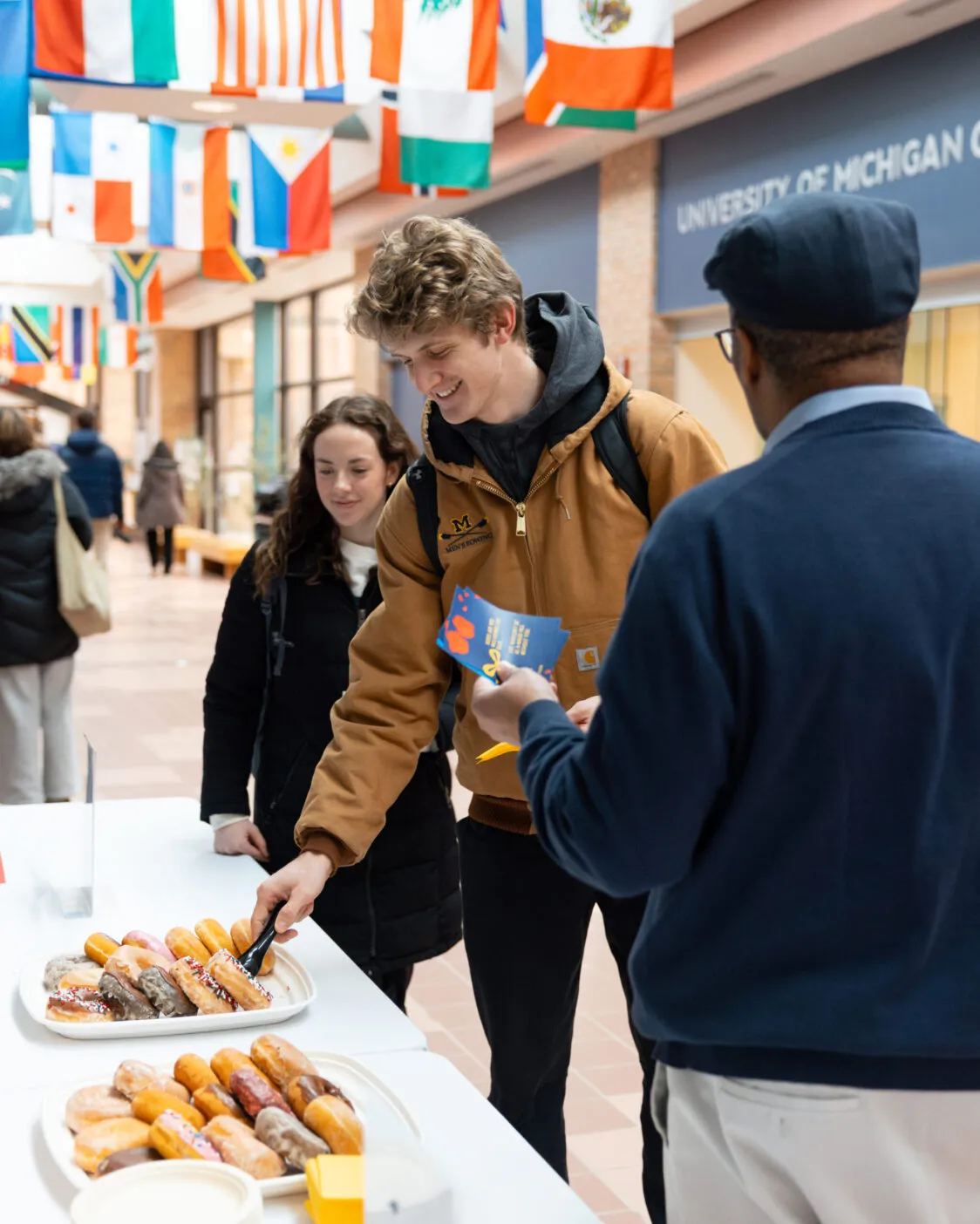 A table of doughnuts is set up in the hallway of Pierpont Commons. Flags from many countries hang from the ceiling. Martino Harmon, Vice President for Student Life, speaks to two students who are selecting doughnuts.