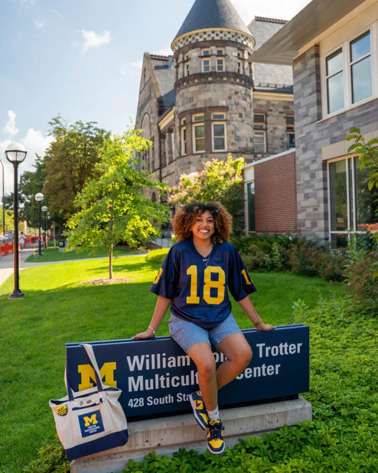 Zoe Wright in a navy University of Michigan jersey with yellow number 18 sits on a campus sign for the William Monroe Trotter Multicultural Center, with a UM tote bag beside them and a stone academic building and green lawn in the background.