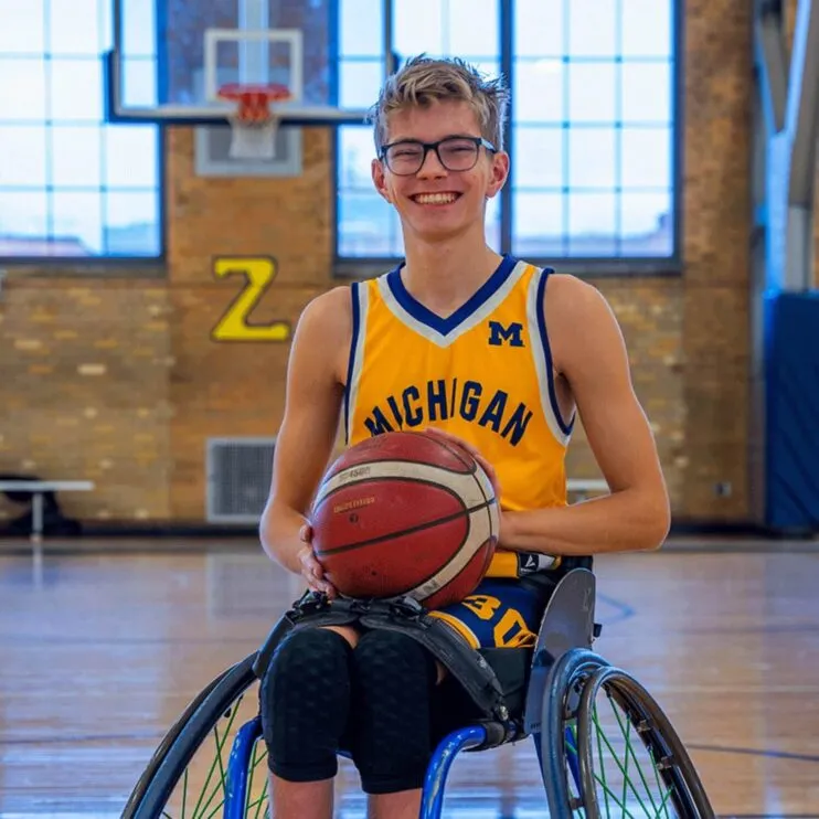 A student with blond hair and glasses sits in a wheelchair, a basketball in his lap. He is wearing a Michigan basketball jersey.