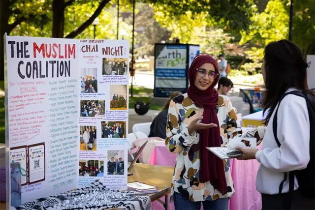 Woman in hijab speaking to a student. A poster board behind her reads: The Muslim Coalition