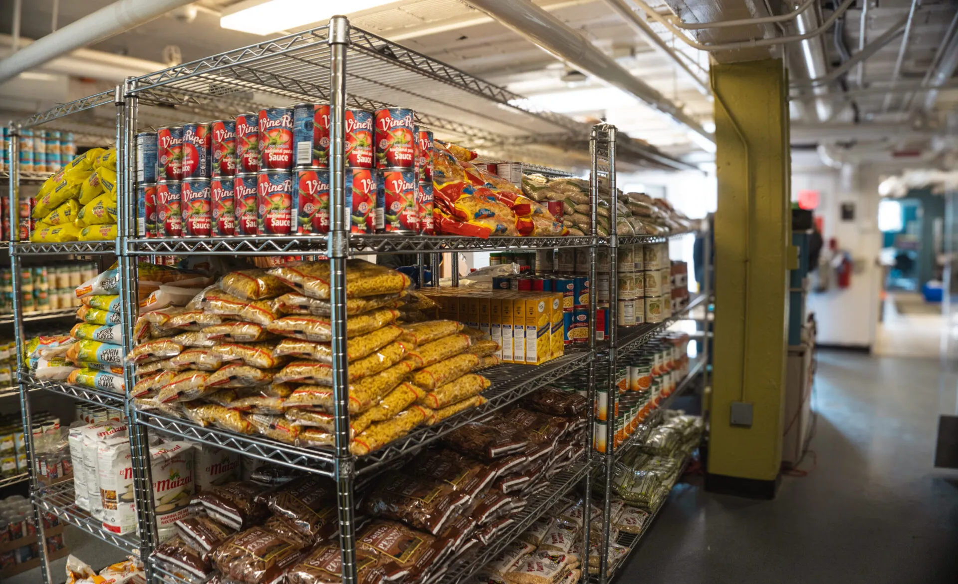A long row of wire shelves hold shelf stable foods like beans, bags of pasta and boxes of mac and cheese.