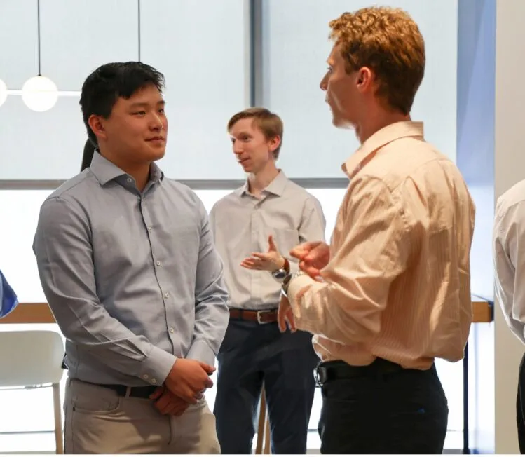 Two male students talk, both dressed in professional clothing. One wears a light blue button down shirt, the other wears a light colored button down shirt.