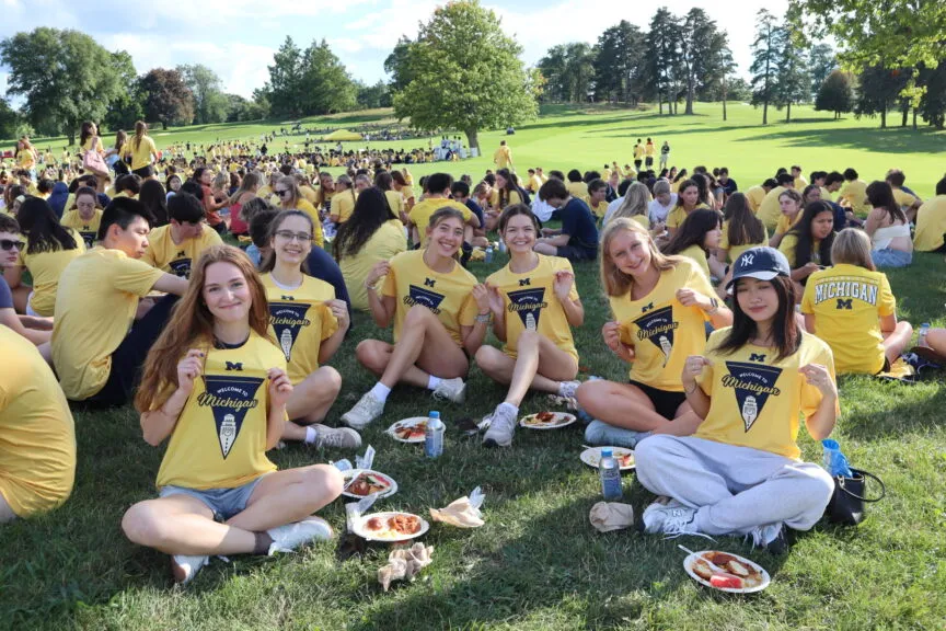 A large group of college students sit on a sunny grassy lawn during an outdoor campus event, many wearing matching yellow “Welcome to Michigan” T-shirts. In the foreground, a group of smiling young women sit cross-legged, holding up their shirts and posing for the camera, with plates of food and drinks on the grass around them. In the background, hundreds more students gather in clusters across a gently sloping green field dotted with trees under a clear sky.