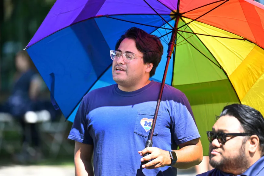 A person in a blue T-shirt with a small U‑M heart logo holds a large rainbow umbrella outdoors.