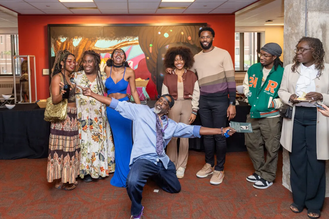 Group of seven adults posing indoors in front of a colorful wall mural; one person kneels in the foreground with arms outstretched holding a pamphlet, while others stand close together in dressy and casual outfits in a lobby-like space.