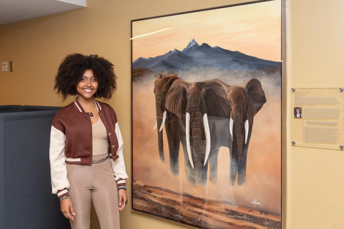 Elsa Olander stands indoors beside a large framed painting of three elephants walking through dusty ground, with a mountain range and warm sunset sky in the background; an informational plaque is mounted on the wall to the right.