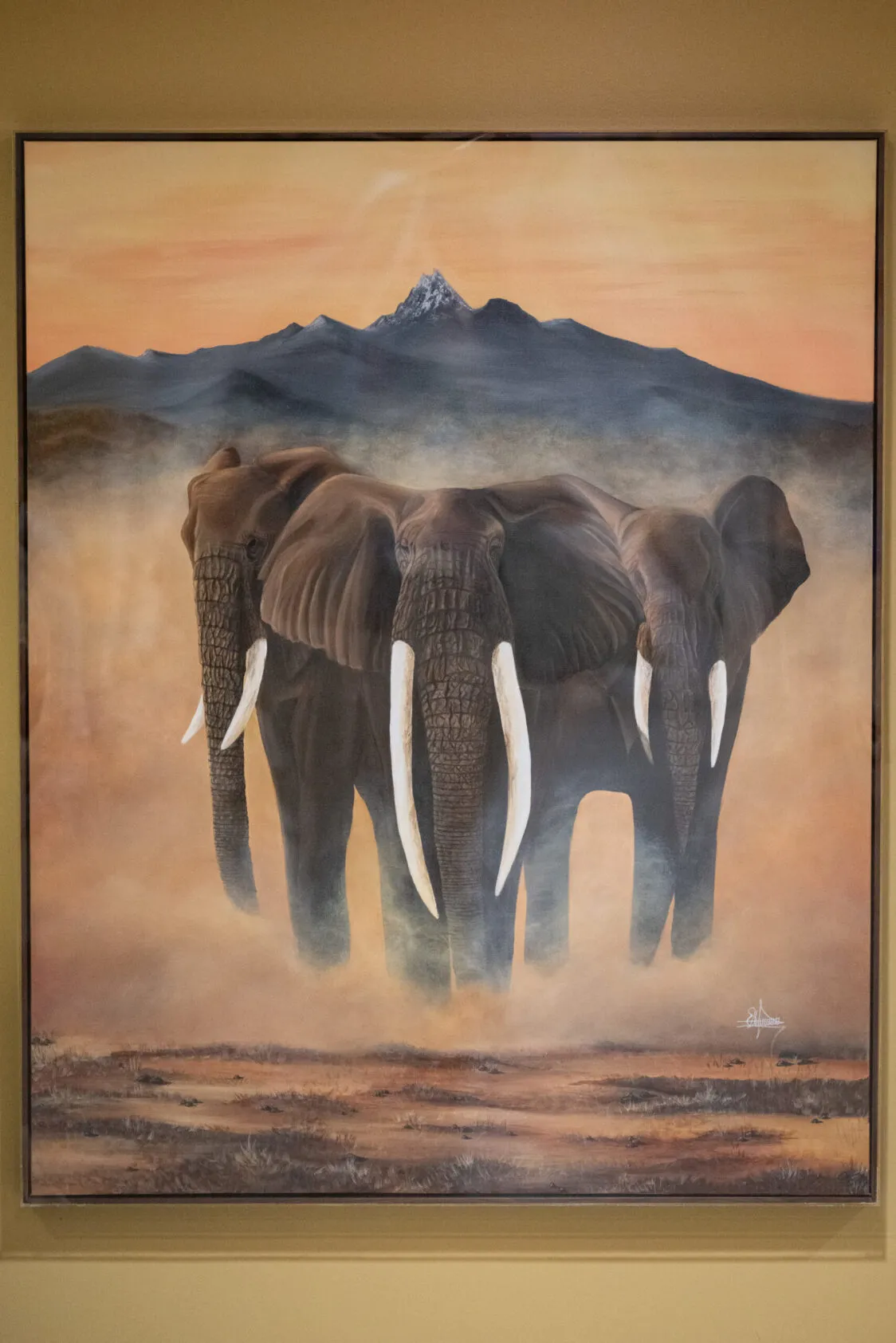 Framed painting of three elephants standing in a cloud of dust, their long tusks prominent, with a dark mountain range and a single snow-capped peak beneath an orange sunset sky.