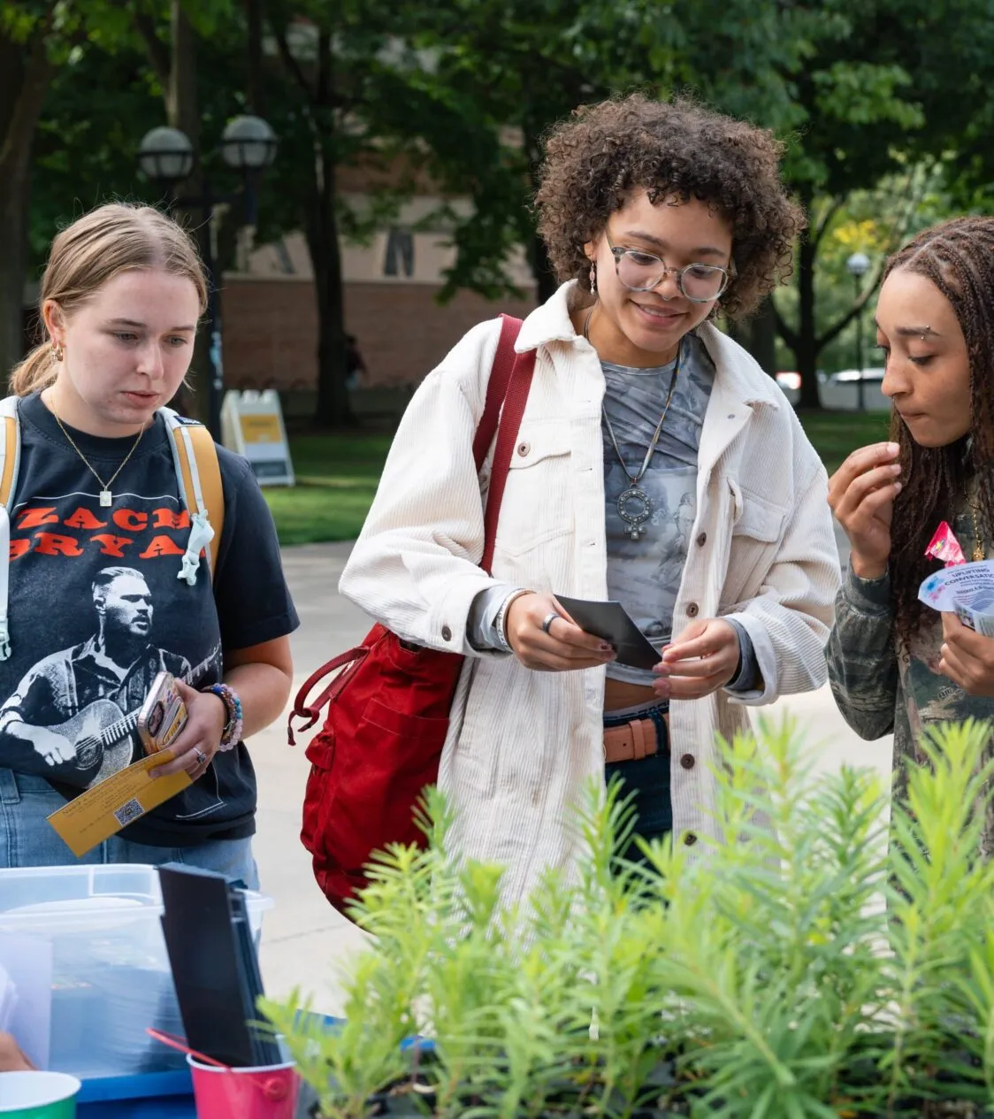 A woman with white skin and brown hair stands next to a woman with light brown skin and brown hair. A third student joins them. She has brown skin and brown hair. They are looking at a flyer.