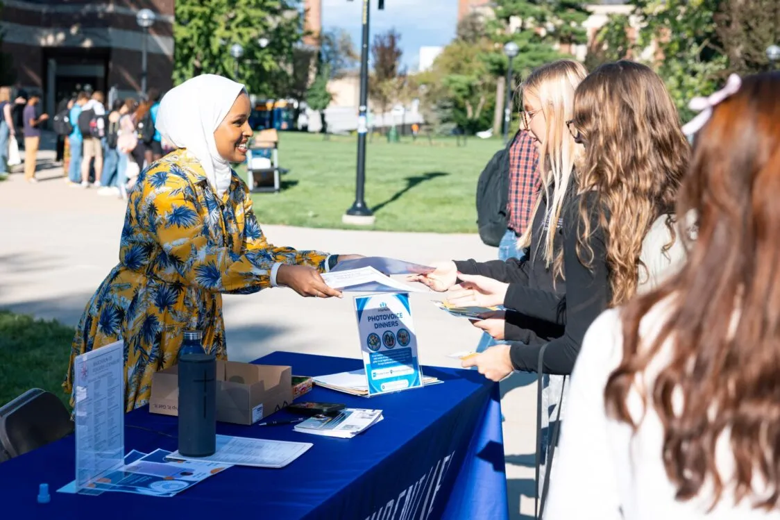 A woman wearing a hijab and a flowered dress stands behind a table. She hands some papers to a group of students.