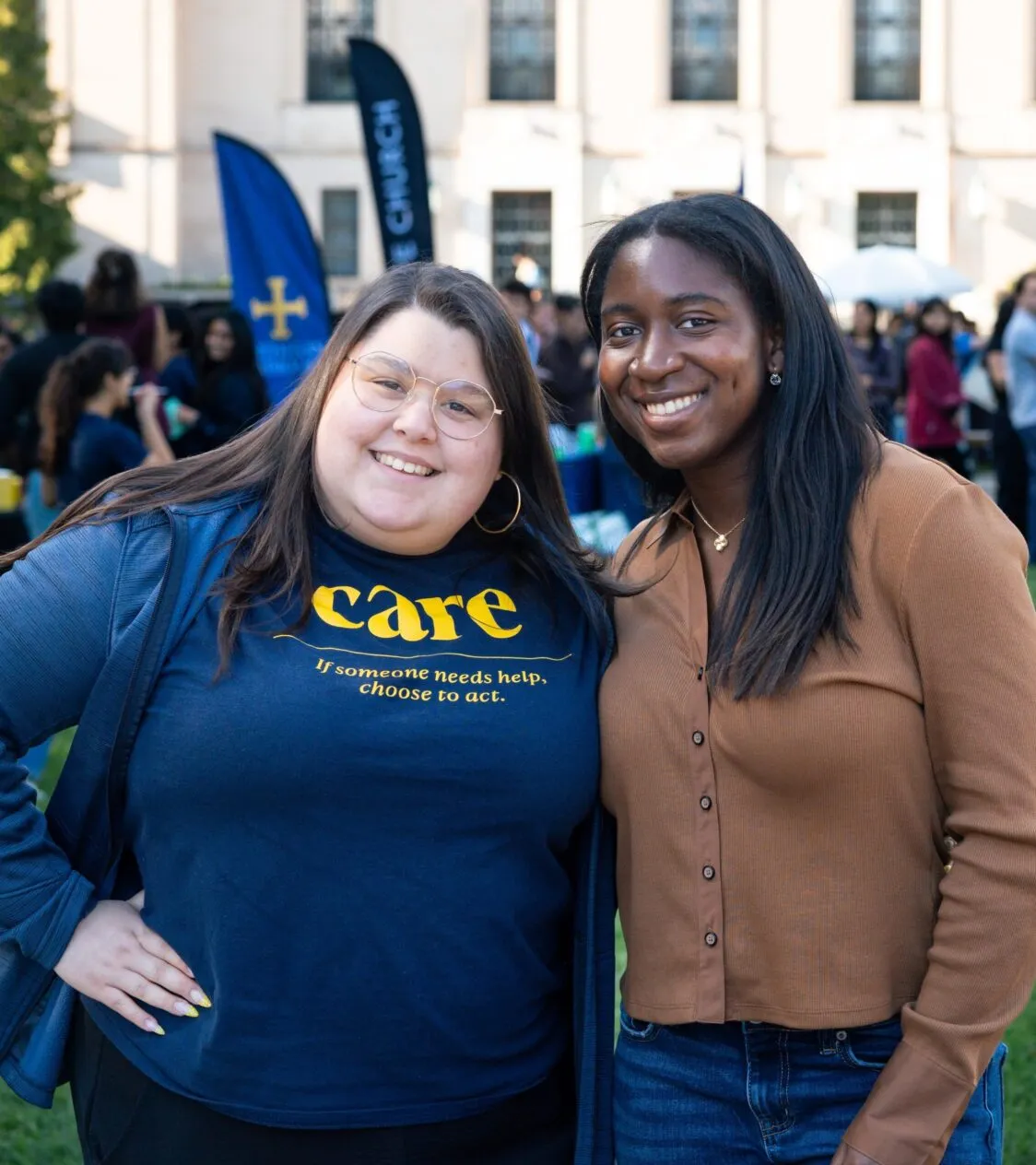 A student with light skin and a blue shirt that reads Care stands with a student with brown skin and a tan shirt. The background is blurred and shows a crowd of people.