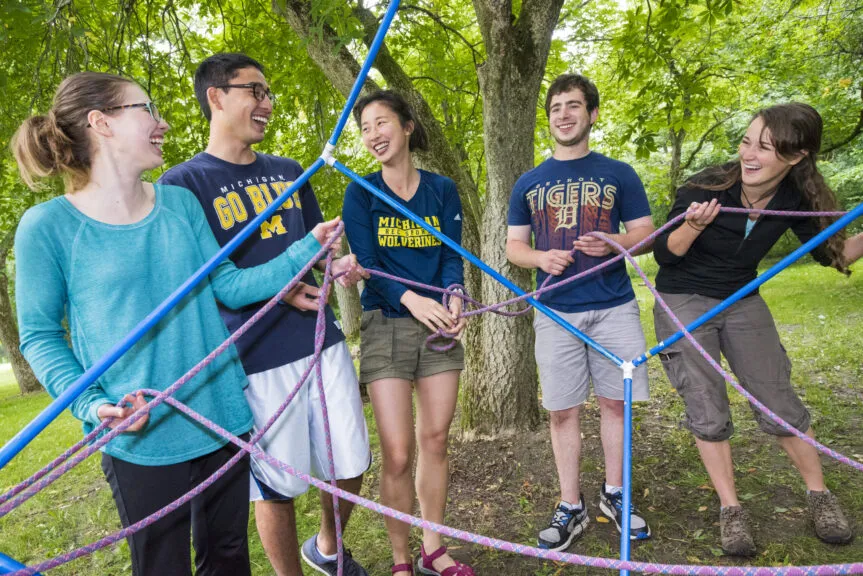 Five people stand outdoors under leafy trees, working together to weave pink ropes through a blue PVC frame as a team-building puzzle; several wear University of Michigan shirts.