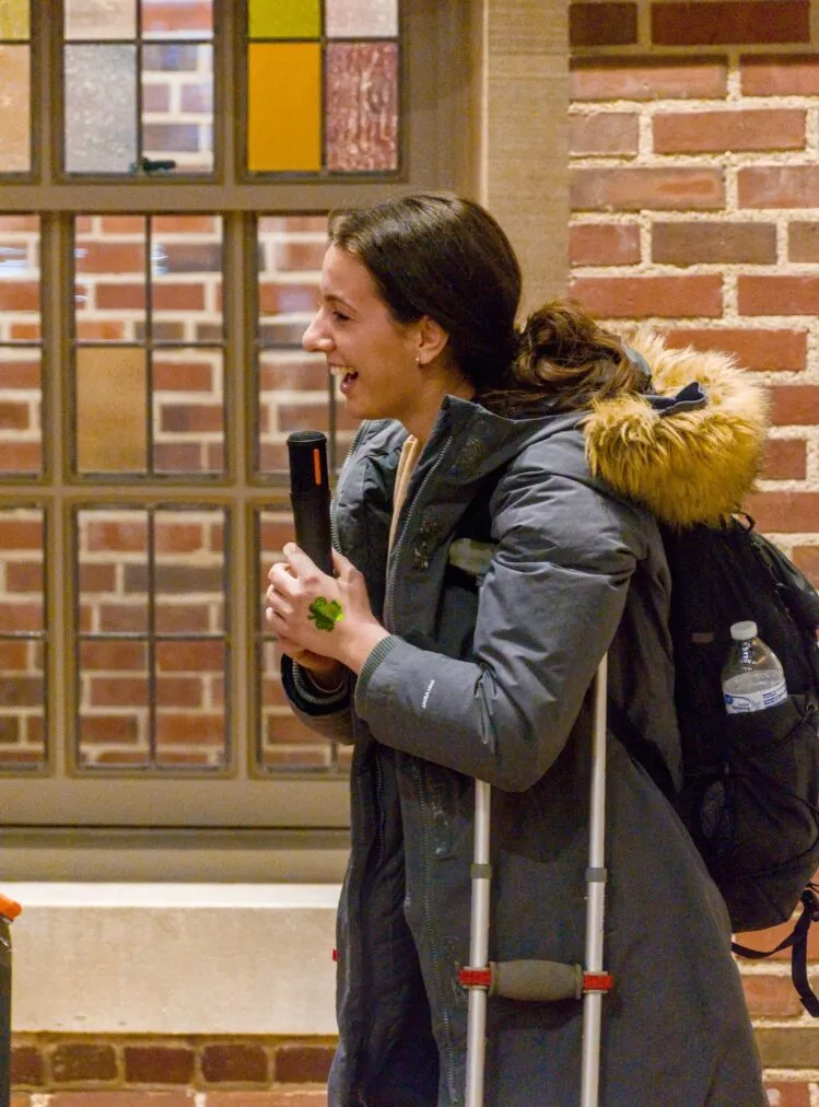 A student holds a microphone. She is laughing and she has a shamrock sticker on her hand. She is wearing a dusty blue parka and leans on a crutch.