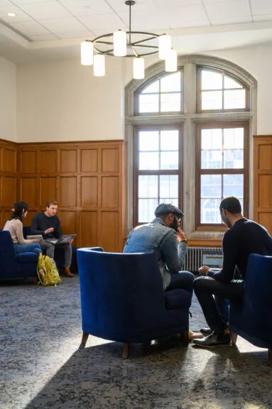Students in discussion sit huddled over a laptop in blue lounge chairs in a wood-paneled study room with tall arched windows; other students work nearby, sunlight streaming across the carpet.